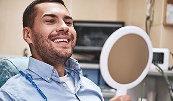 Man with stubble smiling while sitting in dental chair looking at mirror